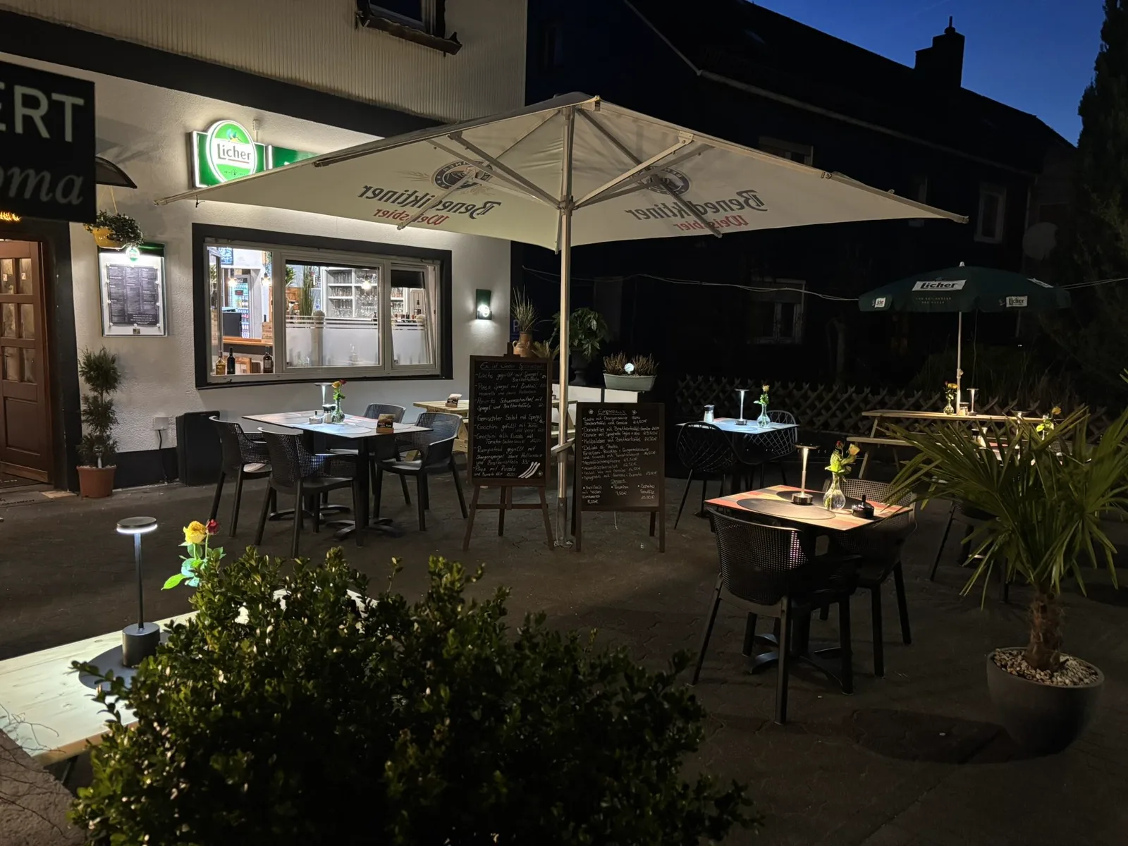 Die Terrasse des Restaurants Pizzeria Roma am Abend: gedeckte Tische unter Sonnenschirmen, beleuchtete Kreidetafeln mit den Tagesempfehlungen